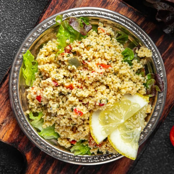 Mediterranean couscous salad with lettuce and lemon wedges in a silver bowl on a wooden board.