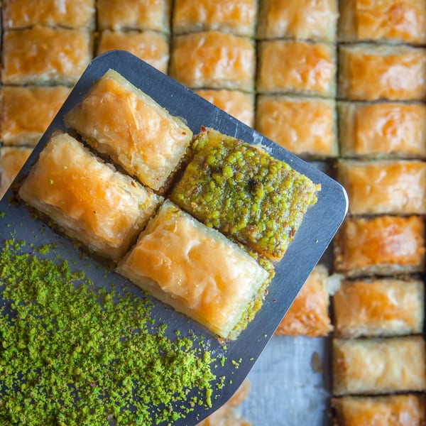 A server lifts a tray of golden baklava, some topped with green pistachios, from a larger pan.