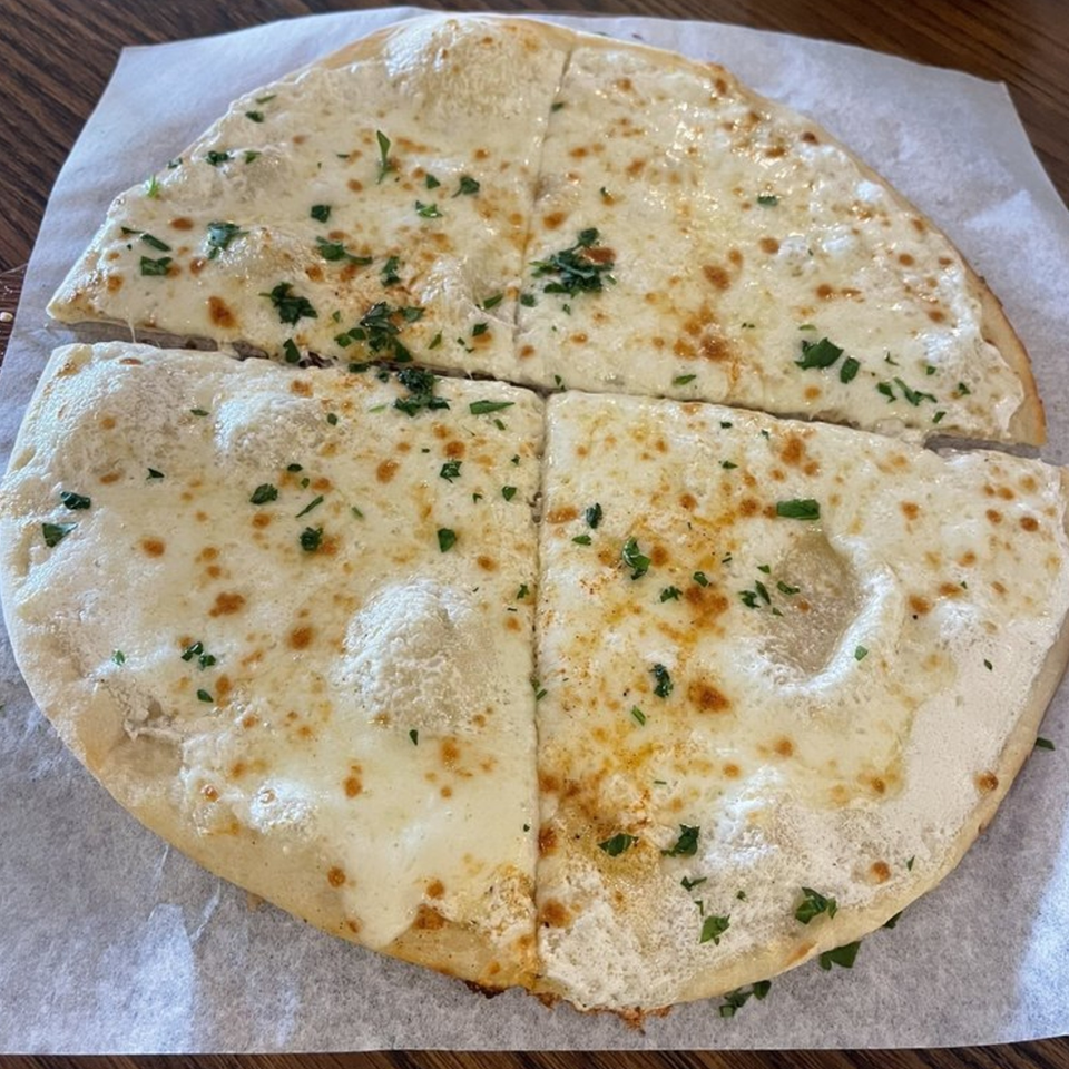 A round, flat Mediterranean bread, topped with melted white cheese and green herbs, cut into four pieces.