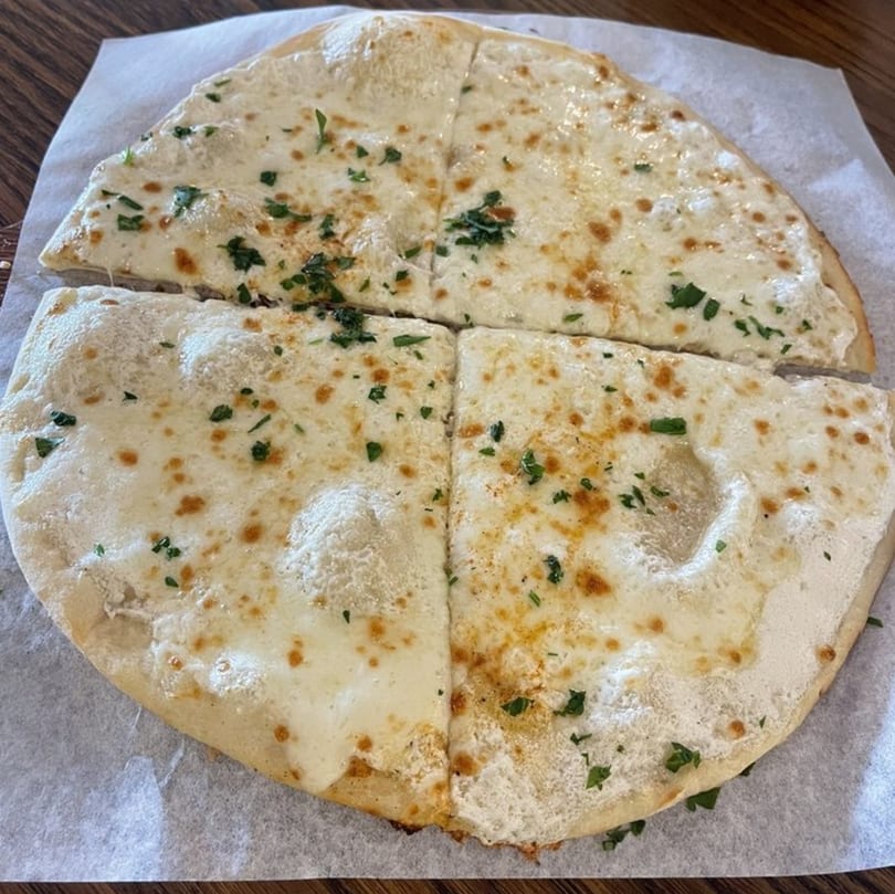 A round, flat Mediterranean bread, topped with melted white cheese and green herbs, cut into four pieces.