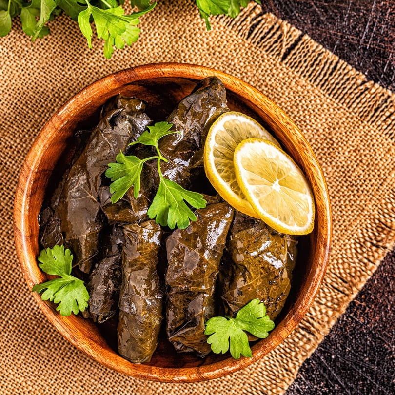 A wooden bowl of stuffed grape leaves (dolmades) garnished with fresh parsley and lemon slices.