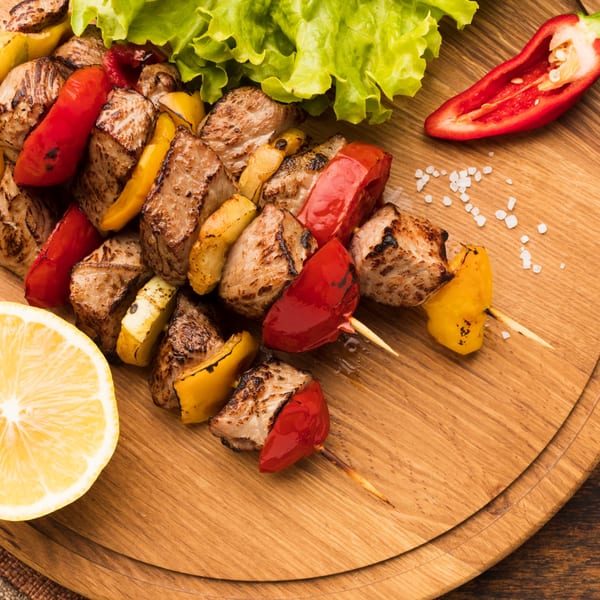 Grilled Mediterranean shish kebabs with red and yellow bell peppers, lemon, and lettuce on a wooden board.